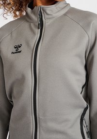 Grå zip-jacka i texturerad tyg, med svart dragkedja och sidofickor. Logotypen är placerad på vänster bröst.