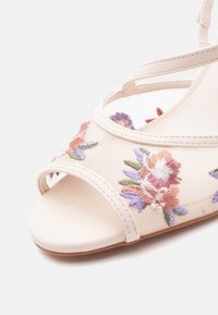 Μπεζ σανδάλι ανοιχτού μπροστινού μέρους με κεντήματα λουλουδιών σε ροζ, μωβ και πράσινο, με λεπτά λουράκια αστραγάλου και λεία σόλα.