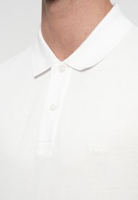 Λευκό πόλο μπλουζάκι με υφή υφάσματος, κλασικό γιακά και κλείσιμο με ένα κουμπί με ανάγλυφο λογότυπο "BOSS" στο στήθος.