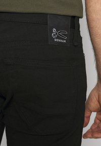 Pantaloni in denim nero con un'apposita etichetta in pelle rettangolare con il testo bianco "DENHAM" e un grafico delle forbici, tasca posteriore standard e dettagli di cucitura.