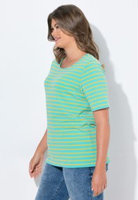 Camisa de manga corta en color verde azulado con rayas horizontales amarillas. Tejido suave, escote redondo, corte relajado, combinada con jeans de mezclilla azules.