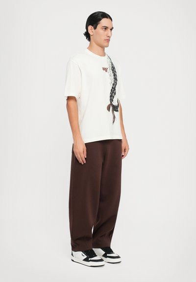 Cream-colored cotton t-shirt with a black hair braid graphic, paired with loose brown trousers and black-and-white sneakers, standing against a plain background.
