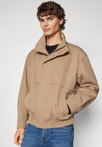 Chaqueta ligera beige con un cuello alto, botones a presión, dos bolsillos frontales y dobladillo elástico, combinada con unos vaqueros.