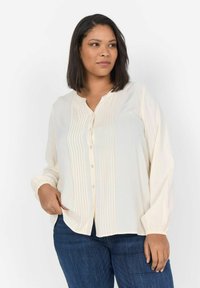 Camicia button-up color crema con frontale plissettato e maniche lunghe con polsini, realizzata in tessuto leggero, indossata con jeans in denim blu scuro.