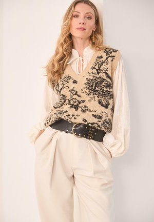 Chaleco de punto beige con patrón floral negro, diseño de escote en V; combinado con pantalones crema de cintura alta y cinturón ancho negro con herrajes dorados.
