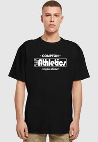 Fekete pamut póló, fehér grafikával ellátott "COMPTON Athletics" felirattal és földgömb mintával; rövid ujjú, laza fazonú.