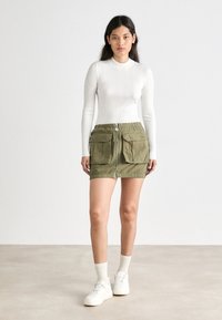 Hvit ribbestrikket langermet topp kombinert med en grønn skjørt med glidelås foran, som har to store lommer og elastisk midje. Modellen har på seg hvite sneakers og sokker.