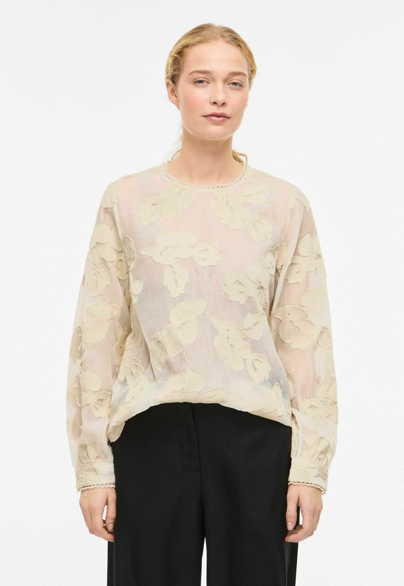 Blouse en voile crème avec broderie florale. Manches longues, encolure ronde avec un bord délicat. Associée à un pantalon noir.