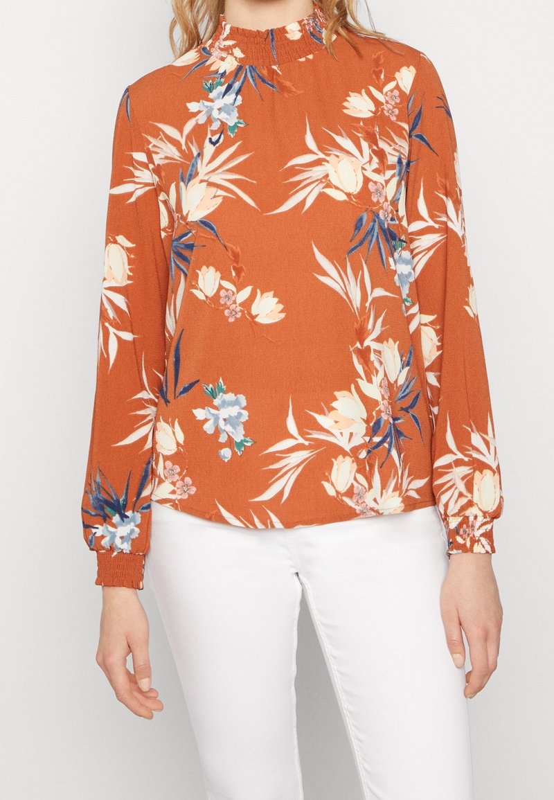 Blusa Color ruggine con motivi floreali in crema e blu, dotata di collo alto e maniche lunghe con polsini elasticizzati. Texture del tessuto morbida.