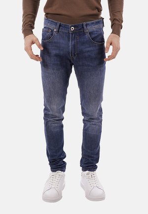 Uomo con jeans blu a sigaretta, maglione marrone e sneakers bianche in piedi davanti a uno sfondo semplice.