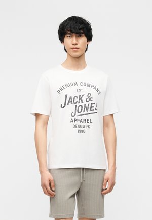 Giovane uomo in piedi che indossa una t-shirt bianca del marchio Jack & Jones e pantaloncini grigio chiaro su uno sfondo chiaro uniforme.
