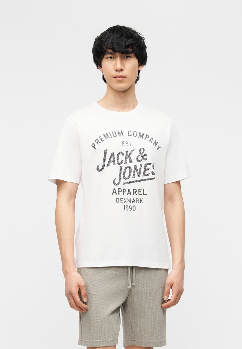 Νέος άνδρας στέκεται φορώντας λευκό T-shirt με το λογότυπο Jack & Jones και ανοιχτό γκρι σορτς σε απλό ανοιχτό φόντο.