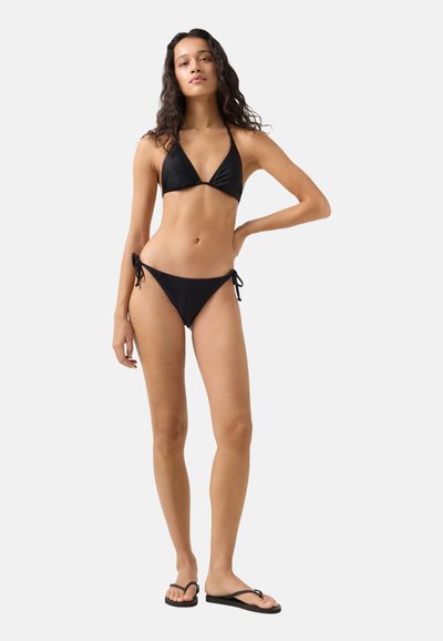 Kvinde stående iført sort bikini i to dele og sorte klipklapper med den ene hånd på hoften mod en ensfarvet baggrund.