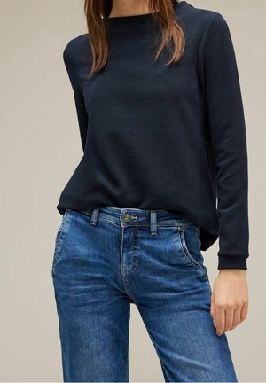 Femme portant un haut noir à manches longues rentré dans un jean bleu en denim, debout avec une main détendue le long du corps.