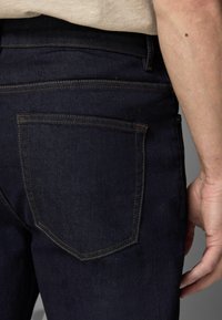 Jeans de mezclilla azul oscuro con un diseño estándar de bolsillo trasero, costuras visibles y una textura suave, parcialmente cubiertos por una camisa.