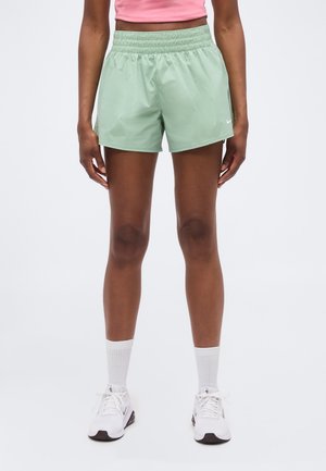 Femeie care poartă pantaloni scurți sport verzi deschis, șosete albe până la gleznă, adidași albi și un top scurt roz pe un fundal simplu.