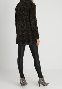 Fuskpälsjacka i mörk olivgrön färg med mjuk textur och ledig passform, kombinerad med glänsande svarta leggings och ankelboots med ormskinnsmönster.