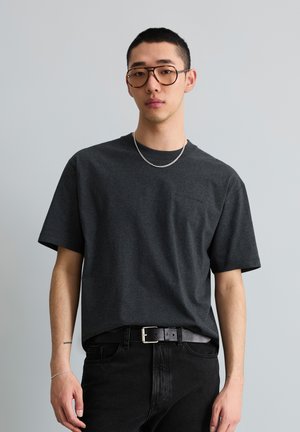 Junger Mann mit kurzen Haaren, der bernsteinfarbene Brillen trägt, dunkelgraues T-Shirt, silberne Halskette und Armband, schwarze Jeans und schwarzen Gürtel vor grauem Hintergrund.