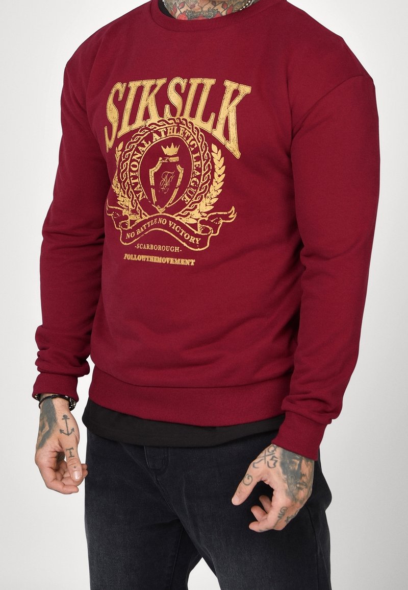 Personne portant une chemise bordeaux à manches longues avec le texte doré "SIKSILK" et un blason, mains tatouées visibles, habillée d'un pantalon noir.