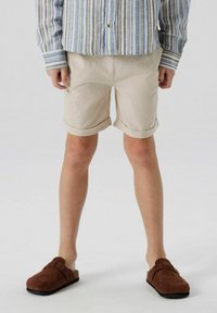 Shorts beige roulés en tissu texturé, associés à des chaussures slip-on en daim marron. La chemise est bleu et blanche à rayures.