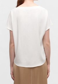 Kortärmad, vit blus med en mjuk yta och rundad nederkant, visad från baksidan, ihop med en beige kjol.