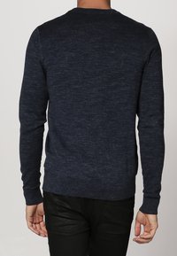 Personne portant un pull à manches longues bleu foncé et un pantalon noir, debout de dos à la caméra devant un fond uni.