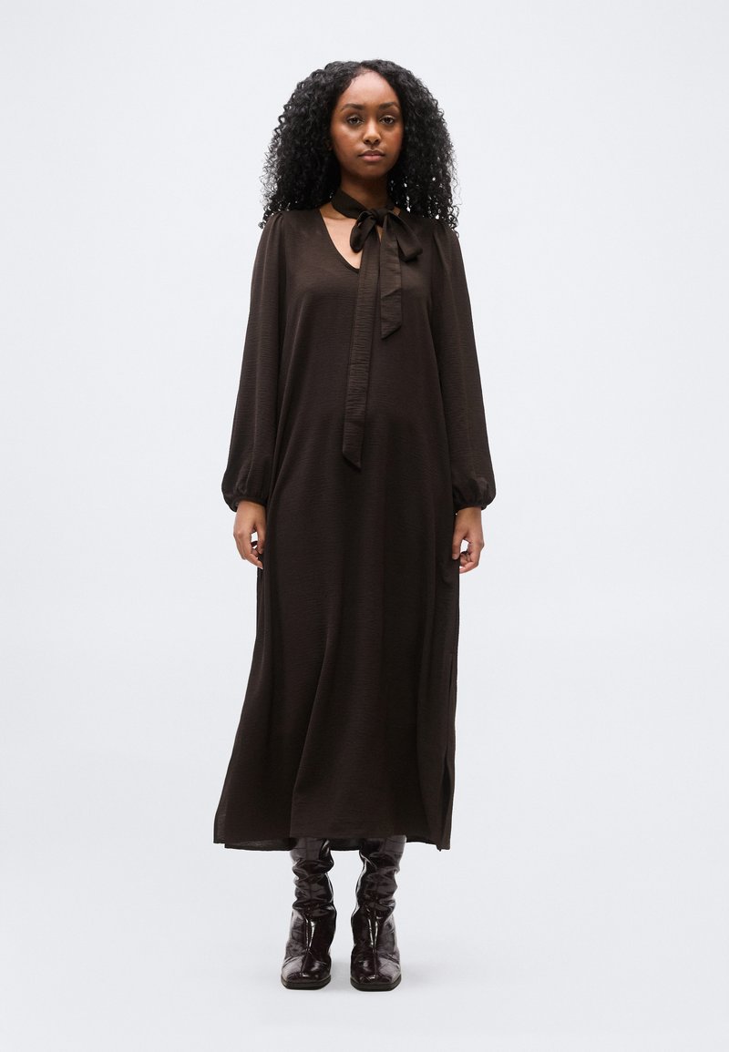 Γυναίκα με σγουρά μαλλιά που φοράει μακρυμάνικο μαύρο φόρεμα maxi με δετή λαιμόκοψη, συνδυασμένο με μαύρα γυαλιστερά μποτάκια αστραγάλου, στέκεται μπροστά σε απλό φόντο.