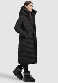 Juodas ilgas pūkinis paltas su aukšta apykakle, siuvinėta struktūra ir užtrauktuko priekyje. Derinamas su juodais šilumą sulaikančiais batais su logotipu.