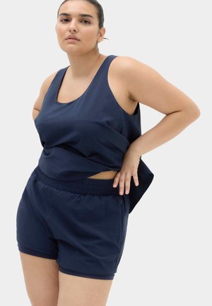 Kvinde iført navyblå ærmeløs top og shorts, poserende med den ene hånd på hoften mod en ensfarvet lys baggrund.