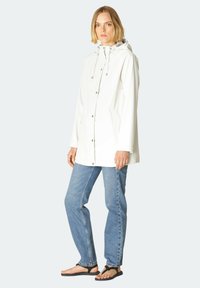 Chaqueta blanca impermeable con capucha, botones en el frente, bolsillos laterales y un corte relajado, combinada con vaqueros azul claro y sandalias negras.