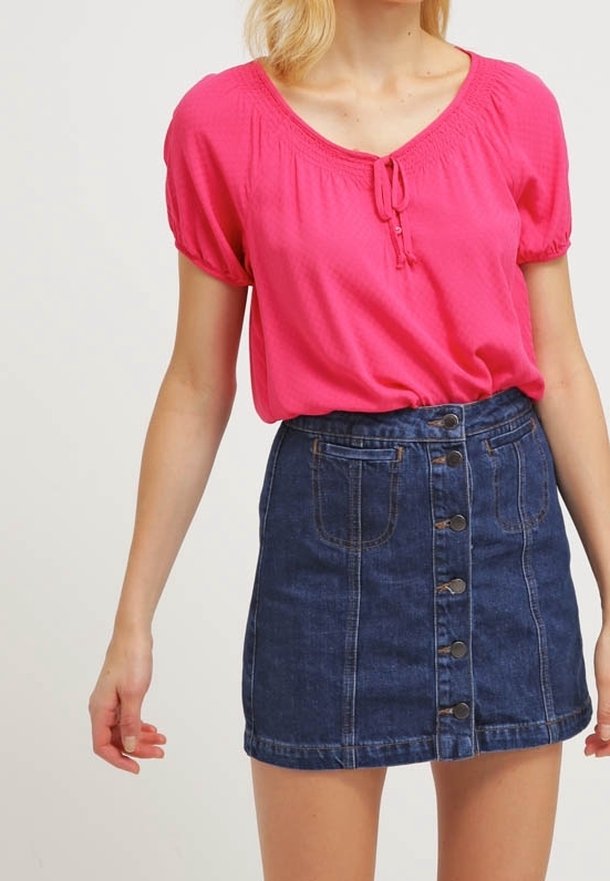 Blouse rose à manches courtes avec un détail noué au niveau de l'encolure, associée à une jupe en denim foncé ajustée avec des boutons et des poches sur le devant.