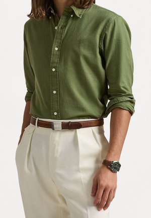 Homme portant une chemise verte à boutons retroussée, un pantalon plissé blanc, une ceinture en cuir marron et une montre avec un bracelet en cuir marron au poignet gauche.