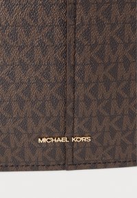 Καφέ συνθετική τσάντα χειρός με ανάγλυφο μοτίβο λογότυπου και χρυσά μεταλλικά στοιχεία. Φέρει την επιγραφή "MICHAEL KORS" σε χρυσά γράμματα στο κάτω μέρος.