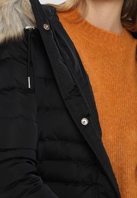 Musta toppatakki, jossa on teksturoitu pinta, turkiksella vuorattu huppu ja hopeanväriset metalliosat. Oranssi neulottu neule näkyy takin alta.