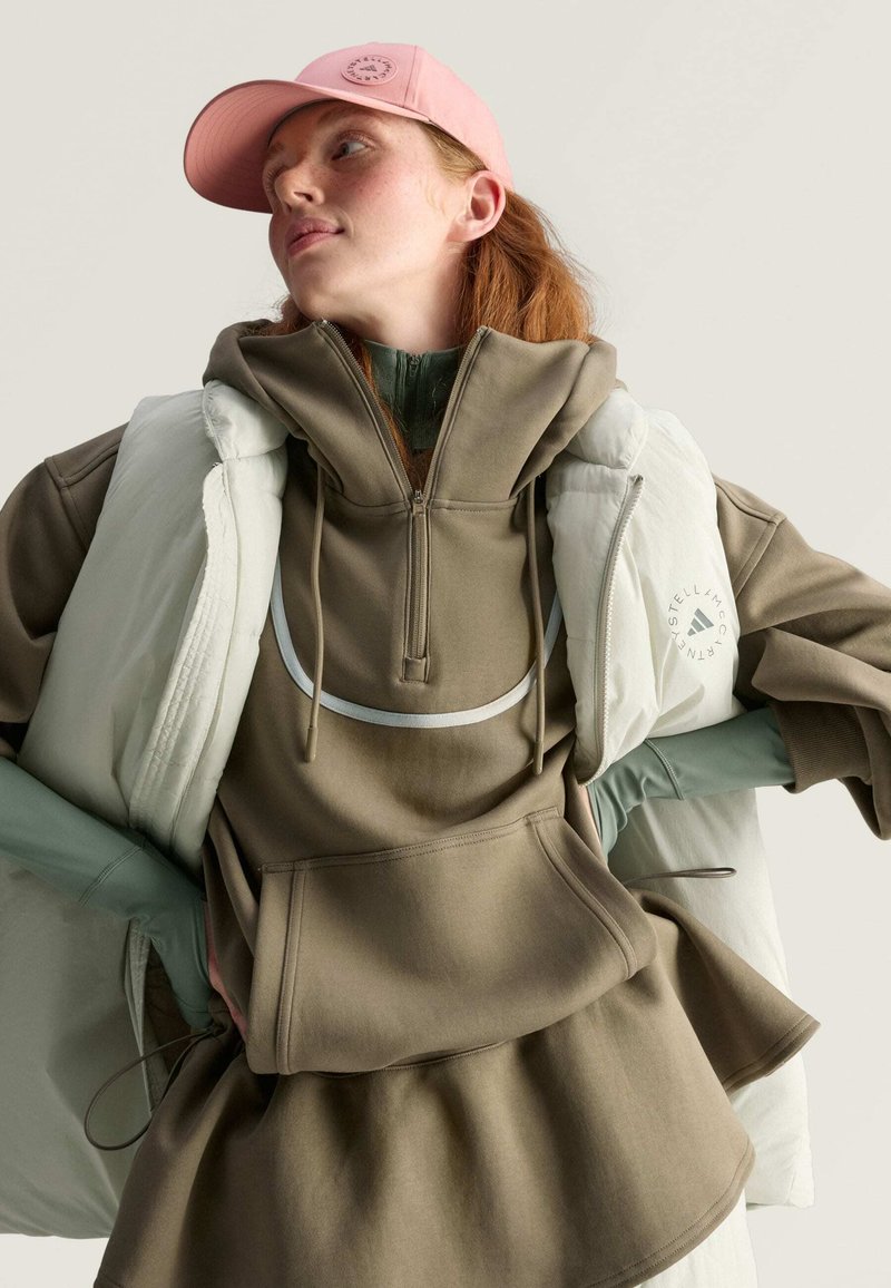 Sudadera verde oliva con cuello con cremallera, llevada bajo un chaleco acolchado blanco. Gorra rosa, con un logo circular, complementa el conjunto.