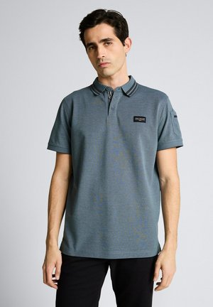 Jeune homme portant un polo à manches courtes bleu-gris avec des rayures noires sur le col, debout devant un fond uni.