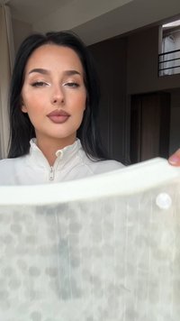 Kobieta z długimi ciemnymi włosami i neutralnym makijażem trzyma półprzezroczysty materiał o subtelnym wzorze w delikatnie oświetlonym pomieszczeniu.