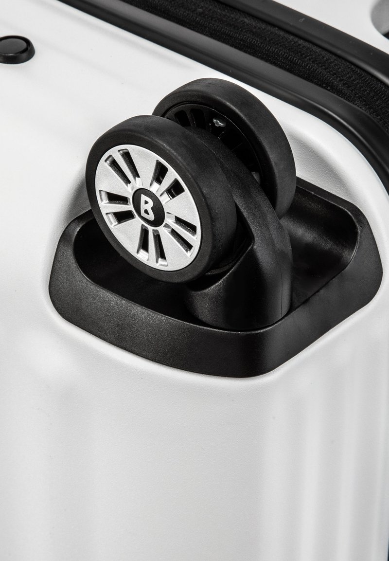 Twarda walizka z błyszczącej białej powierzchni, z podwójnymi czarnymi gumowymi kółkami na czarnej podstawie oraz srebrnym wzorem kółek z centralnym logo.