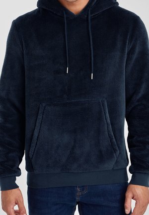 Man wearing a navy blue plush hoodie with front pocket and drawstrings, paired with dark blue jeans.