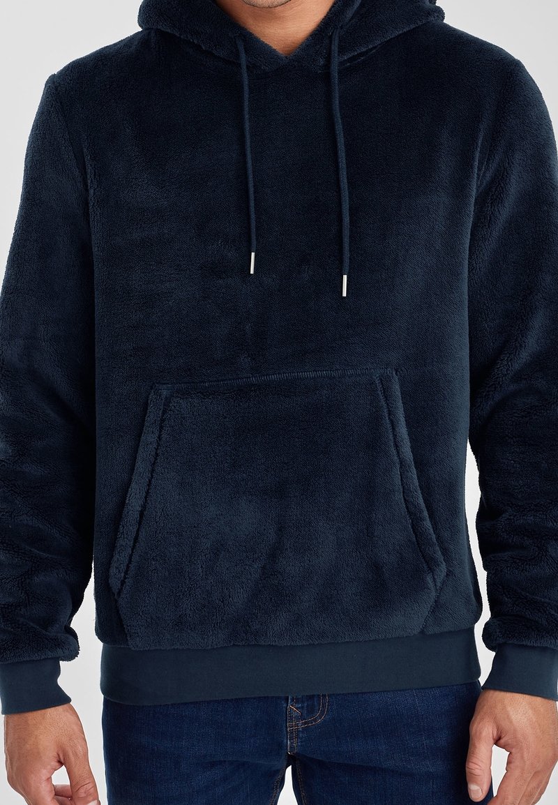 Homme portant un sweat à capuche en peluche bleu marine avec poche avant et cordons, associé à un jean bleu foncé.