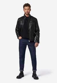 Zwarte leren jas met twee ritszakken op de borst, gedragen over een zwart shirt, gecombineerd met donkerblauwe jeans en zwarte enkellaarsjes.