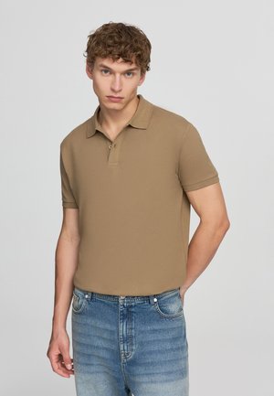 Giovane uomo con capelli ricci che indossa una polo a maniche corte color beige e jeans blu sbiaditi, su uno sfondo chiaro e semplice.