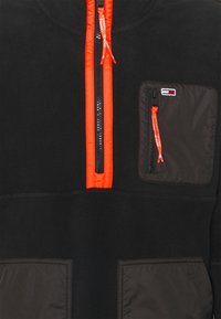Black fleece jacket with bright orange zipper trim and pull tabs, featuring a zippered chest pocket with a small flag logo.