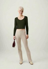 Top cropped de manga longa verde oliva e calças beiges de cintura alta com textura em ribas, combinados com sapatos de salto beiges e uma pequena mala escura.