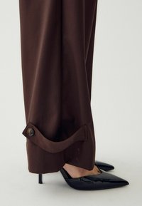 Smeđe hlače s prevrnutim rubom i detaljem na dugmetu, uparene s crnim cipelama na visoku petu s šiljastim vrhom, koje prikazuju glatku, sjajnu teksturu.