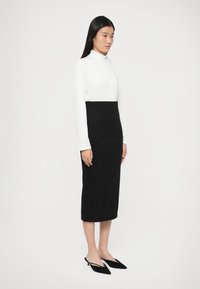 Μαύρη φούστα με εφαρμοστή σιλουέτα, συνδυασμένη με λευκό ζιβάγκο μακρυμάνικο. Το σύνολο διαθέτει απαλές υφές και μυτερές μαύρες παπούτσια.