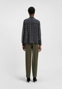 Blusa a maniche lunghe in tessuto trasparente e decorato, con scollatura arricciata e polsini elasticizzati, abbinata a pantaloni sartoriali verde oliva e tacchi neri.