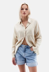 Světlý béžový košilový top s texturovanou tkaninou a náprsní kapsou, v kombinaci s ohrnutými modrými denimy. Jednoduchý, casual outfit.
