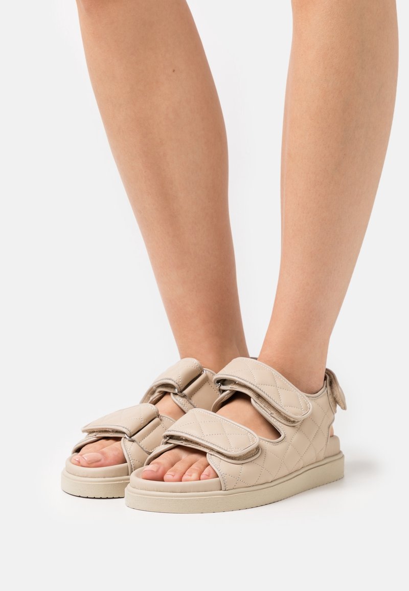 Jambes portant des sandales matelassées beiges avec deux larges bandes Velcro, orteils ouverts et semelle plate sur fond blanc.