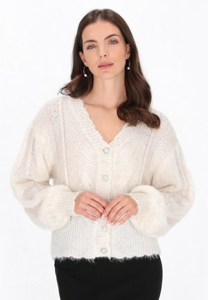Vrouw met golvend bruin haar draagt parel oorringen en een donzige witte gebuttonde cardigan, staand met handen gevouwen bij de taille.
